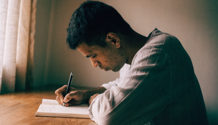 Homem escrevendo em um caderno aberto sobre a mesa, concentrado, em um quarto simples
